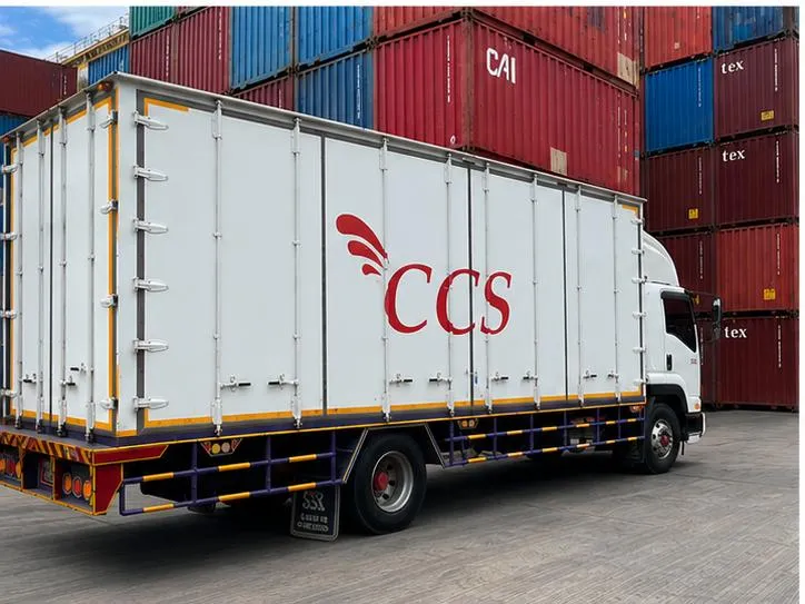 A white CCS box truck parked in front of stacked shipping containers